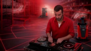 DJ stoji v skladišču za mešalno mizo, v ozadju pa so avtomatizirani viličarji podjetja Linde Material Handling.