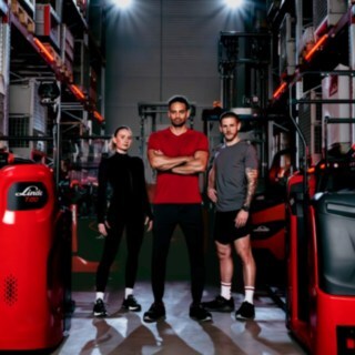 Ženska in dva moška v športnih oblačilih stojijo v skladišču med viličarji podjetja Linde Material Handling.